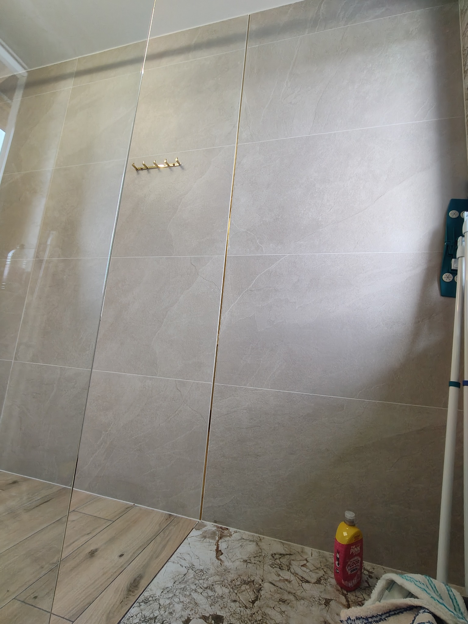 Beżowe, marmurowe płytki w łazience z złotymi akcentami, widoczne wieszaki na ścianie, mop i płyn do mycia podłóg. Ukończona realizacja glazurnicza.