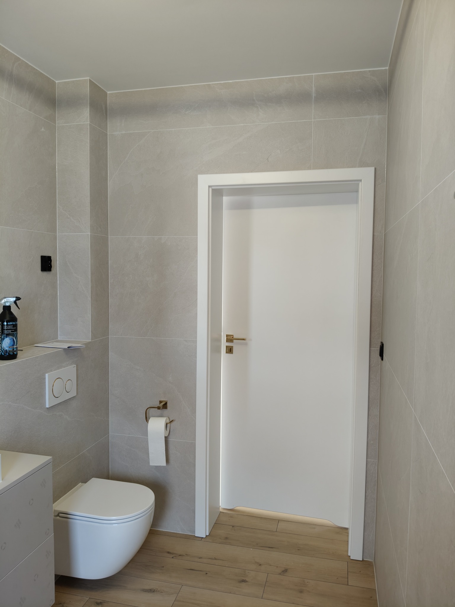 Nowoczesna łazienka po remoncie: szare płytki imitujące kamień, biała wisząca toaleta i drzwi z złotą klamką, drewnopodobna podłoga, minimalistyczny design.