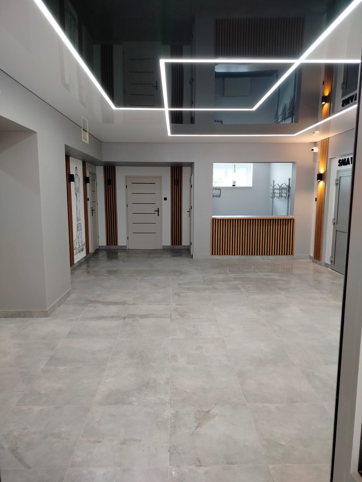 Wykończone wnętrze biura z błyszczącym sufitem, oświetleniem LED, szarymi ścianami, drewnianymi panelami i jasnymi płytkami podłogowymi. Widok na recepcję.