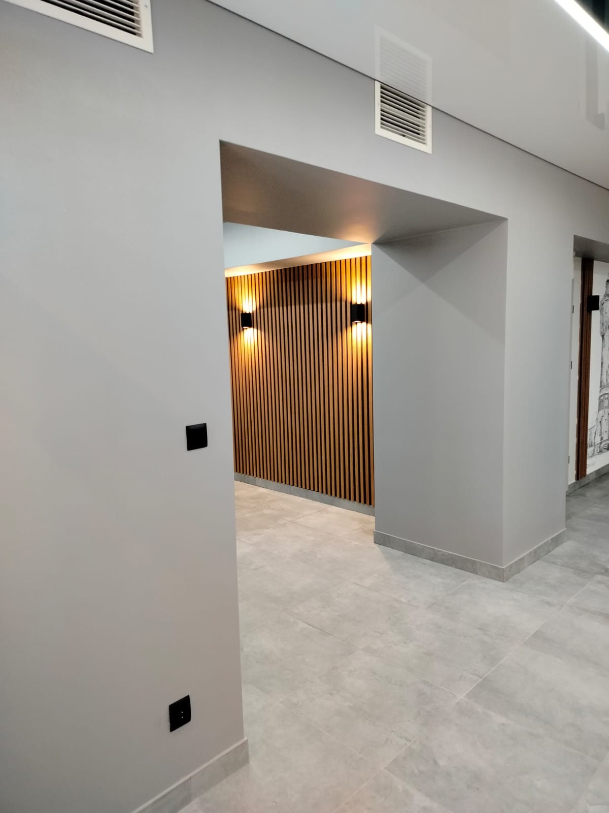 Nowoczesne wnętrze biurowe z drewnianą ścianą lamelową i szarą kolorystyką ścian. Widoczne detale wykończeniowe i oświetlenie.