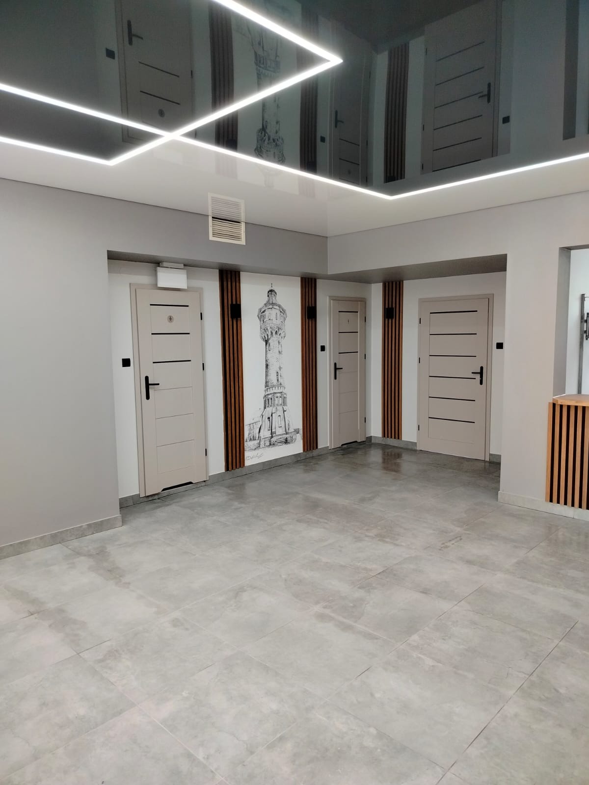 Jasne wnętrze biurowe z szarymi płytkami podłogowymi, beżowymi drzwiami z czarnymi klamkami i lustrzanym sufitem z oświetleniem LED. Na ścianie grafika wieży.