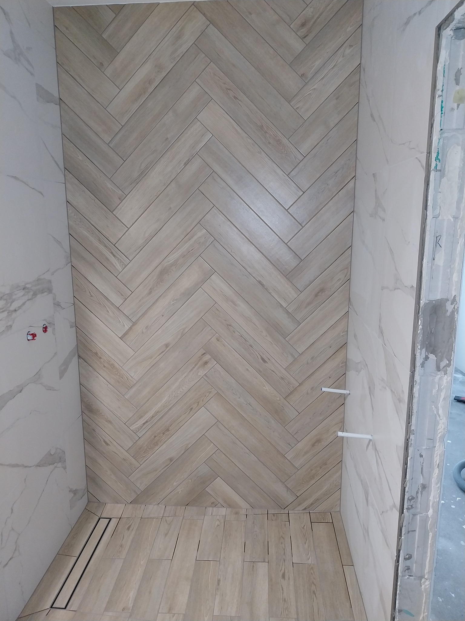 Wykończona kabina prysznicowa z beżowymi płytkami ułożonymi w jodełkę na ścianie i podłodze. Białe płytki imitujące marmur na bocznych ścianach. Widoczny odpływ liniowy.