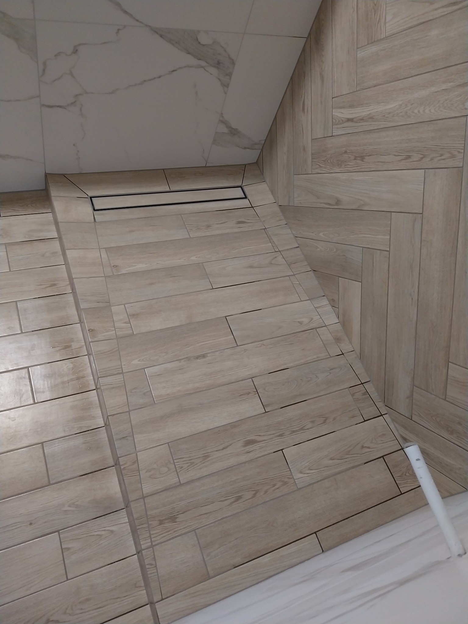 Nowoczesna kabina prysznicowa z odpływem liniowym, wykończona płytkami imitującymi drewno w odcieniach beżu i szarości. Ułożenie płytek w jodełkę na ścianie.