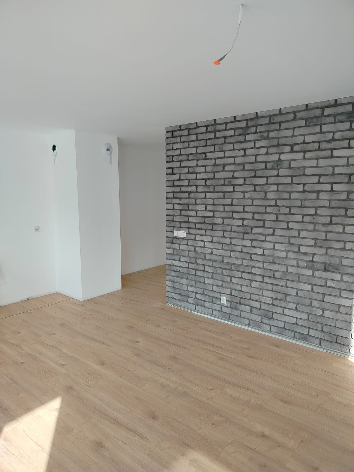 Jasne panele podłogowe i ściana z szarej cegły dekoracyjnej w nowocześnie wykończonym wnętrzu. Widoczne gniazdka elektryczne i wystające kable oświetleniowe.