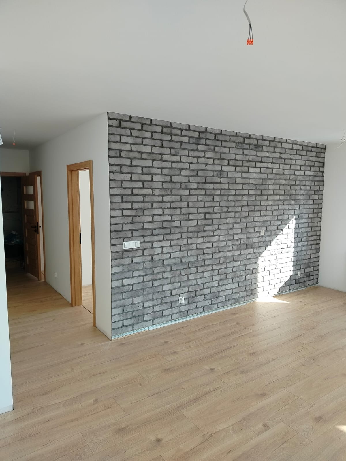 Pokój po metamorfozie: ściana z szarej cegły dekoracyjnej, jasne panele podłogowe, białe ściany. Widoczne gniazdka elektryczne i fragment instalacji pod sufitem.