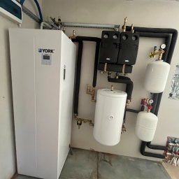 SYLWESTER SORDYL ECOINSTAL - Biała jednostka wewnętrzna z logo YORK podłączona do instalacji hydraulicznej z czarnymi rurami, zbiornikami wyrównawczymi i manometrami, zamontowana w kotłowni z surowym betonowym sufitem.