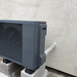 SYLWESTER SORDYL ECOINSTAL - Szara jednostka zewnętrzna urządzenia grzewczego, umieszczona na betonowych bloczkach, z podłączonym przewodem owiniętym srebrną taśmą, na tle surowej, jasnej ściany budynku.