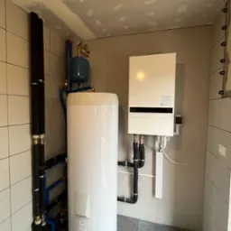 Wnętrze kotłowni z zamontowanym białym zasobnikiem ciepłej wody, niebieskim zbiornikiem wyrównawczym, oraz wiszącym białym kotłem grzewczym na ścianie z szarych płytek. Rury w izolacji termicznej...
