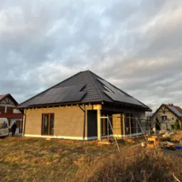 Dom jednorodzinny w trakcie budowy z zamontowanymi panelami słonecznymi na dachu pokrytym dachówką, ocieplony styropianem, widoczne rusztowania i materiały budowlane na działce, pochmurne niebo w tle.