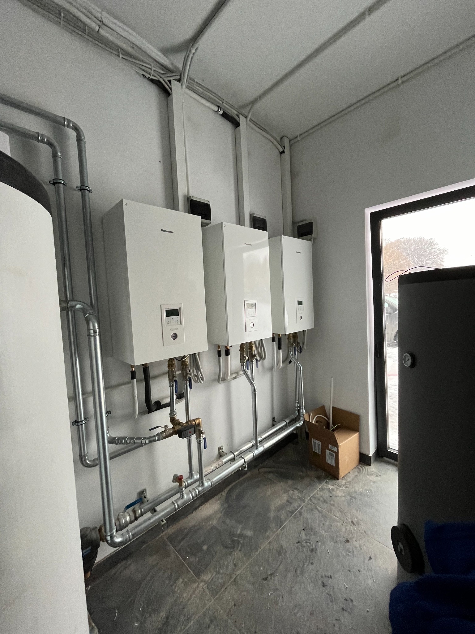 Trzy białe jednostki grzewcze Panasonic zamontowane na ścianie w pomieszczeniu technicznym, z widocznymi rurami i instalacją hydrauliczną, karton leżący na podłodze, widok na zewnątrz przez okno.