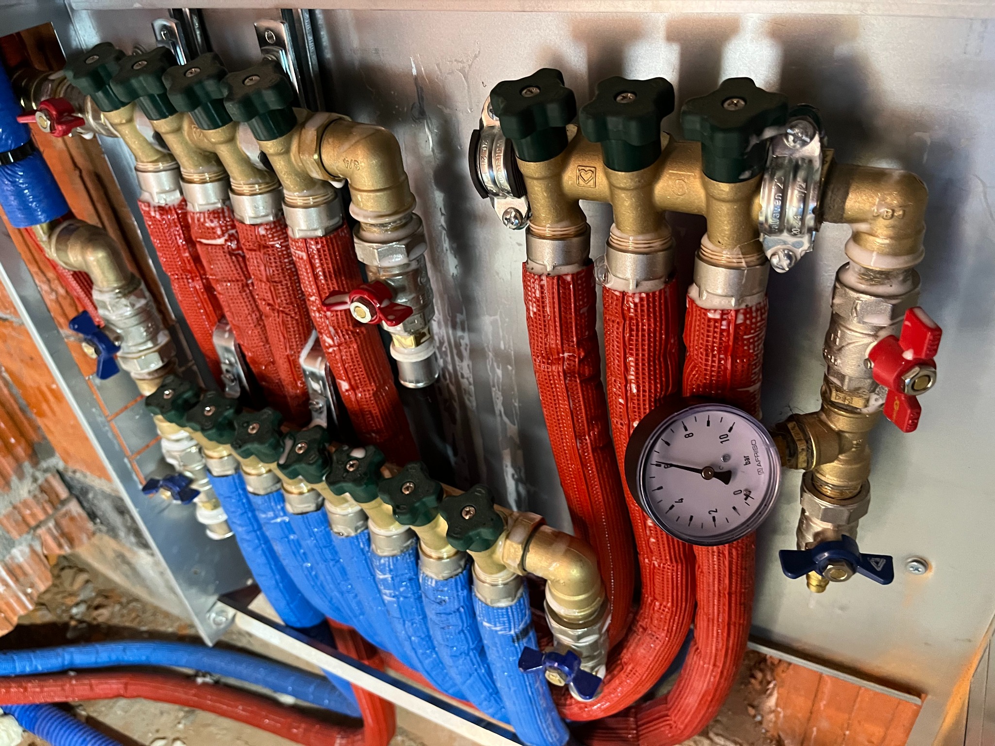 Rozdzielacz instalacji grzewczej z rurami w izolacji, manometrem i zaworami, widok z góry pod kątem.