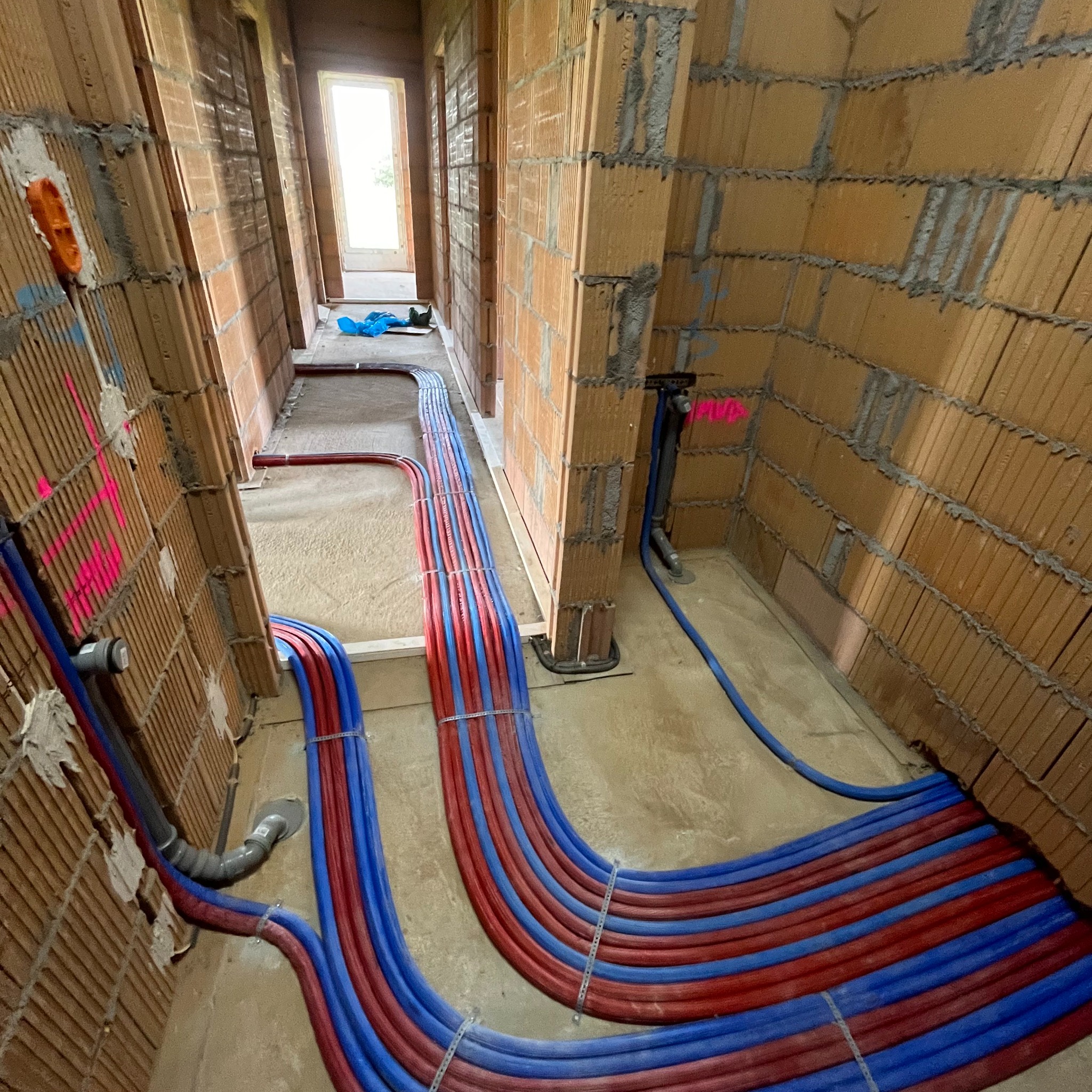 Instalacja rur grzewczych podłogowych w surowym stanie, czerwono-niebieskie rury prowadzone wzdłuż ścian z cegły, widoczny rozdzielacz i elementy kanalizacji.