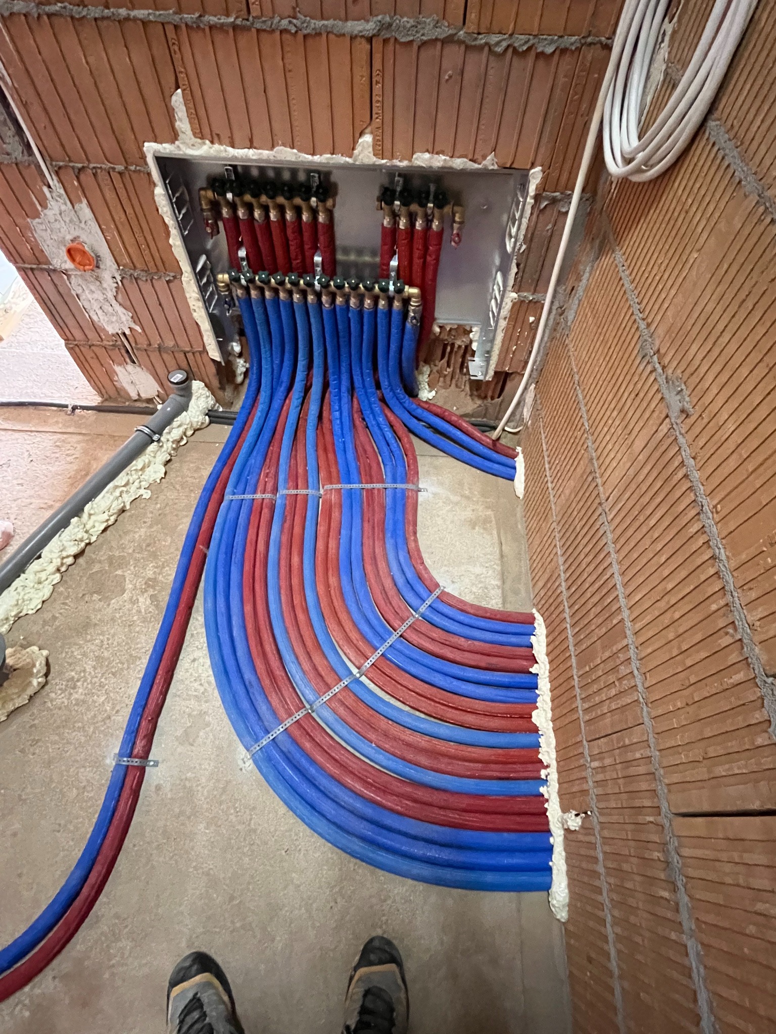 Rozdzielacz ogrzewania podłogowego z podłączonymi rurami w kolorach czerwonym i niebieskim, zamontowany w ścianie z cegły, widoczne mocowania rur i pianka montażowa, ujęcie z dołu z fragmentem obuwia.