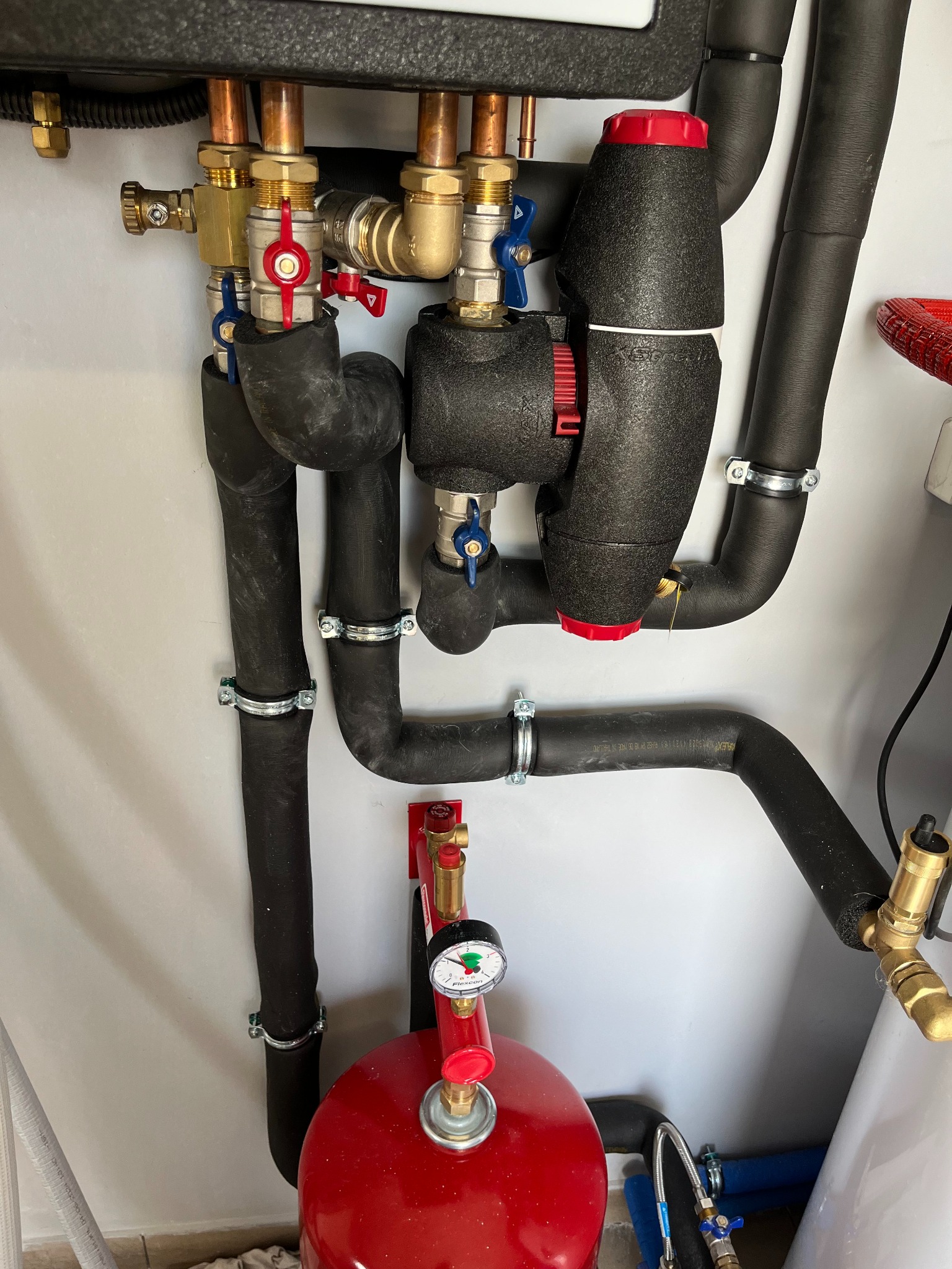 Instalacja hydrauliczna z rurami w izolacji termicznej, zaworami i manometrem, zamontowana na jasnej ścianie.