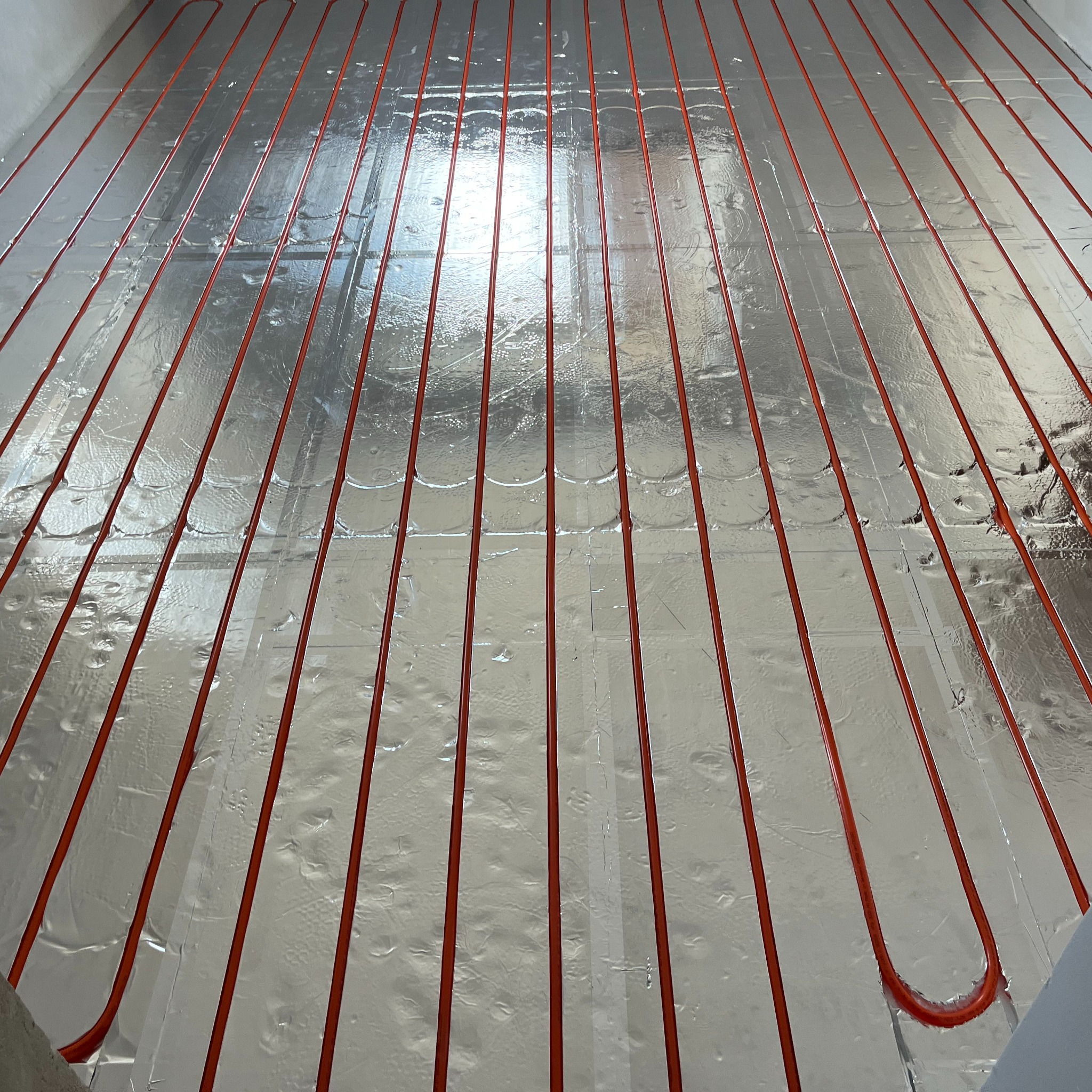 Czerwone rurki ogrzewania podłogowego ułożone na srebrnej folii izolacyjnej, widok z góry, instalacja w trakcie montażu.