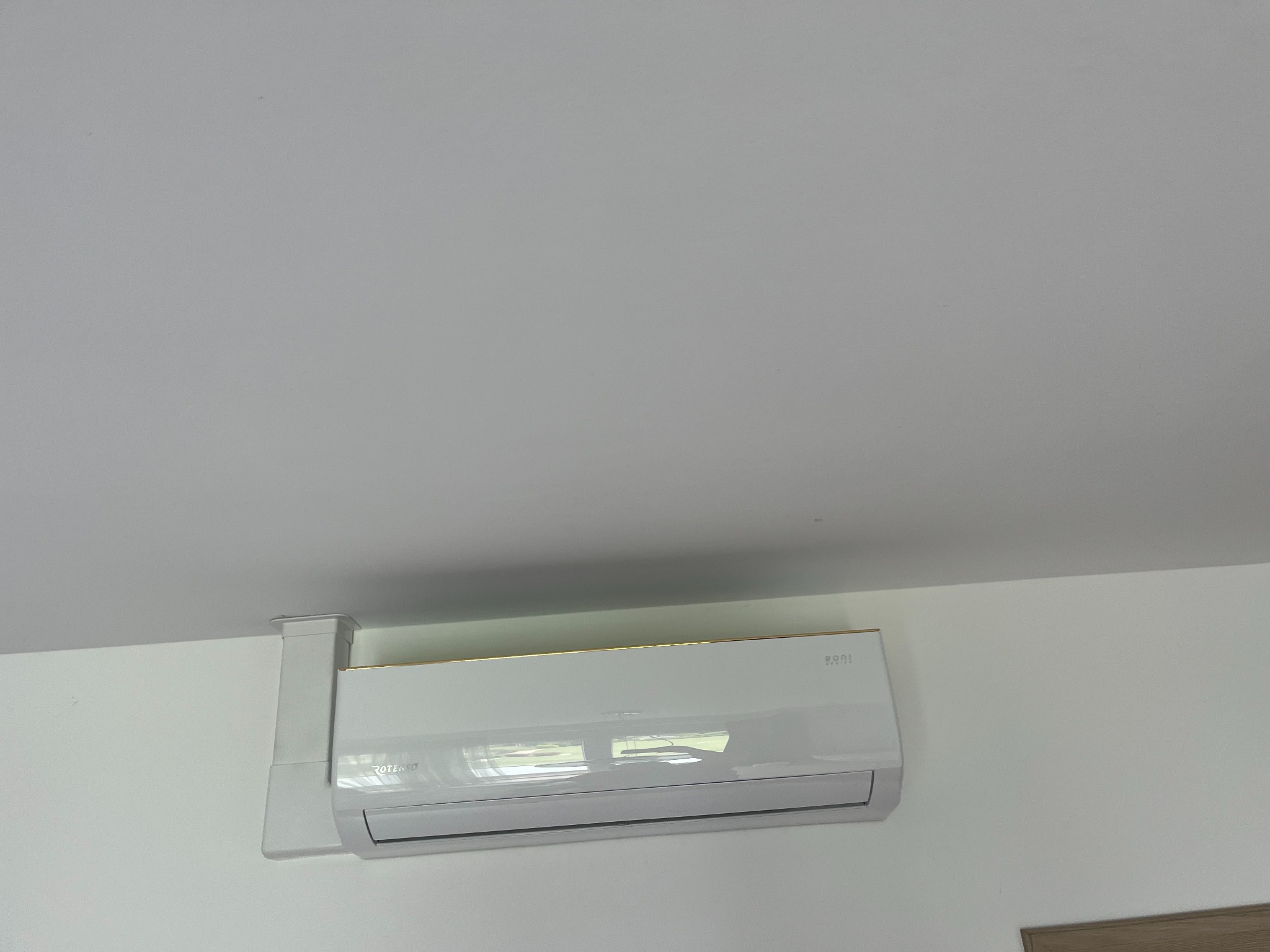 Biała jednostka klimatyzacji z złotym paskiem u góry zamontowana na białej ścianie, widoczne elementy instalacji.