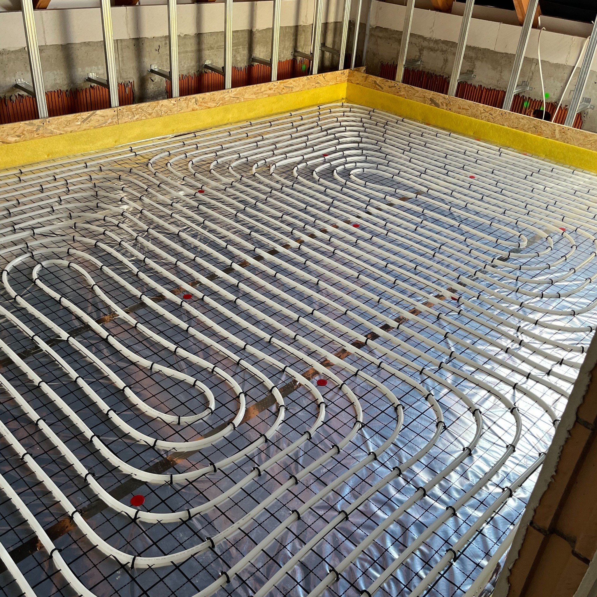 Białe rury ogrzewania podłogowego ułożone w pętle na folii izolacyjnej z czarną siatką, zamocowane czarnymi klipsami, widoczne czerwone znaczniki, ściany z metalowymi profilami i żółtą izolacją...