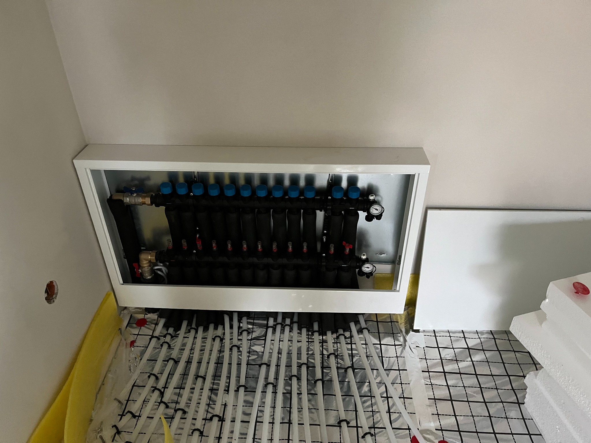 Rozdzielacz ogrzewania podłogowego w białej obudowie, rury ułożone na folii z rastrem, widoczne elementy instalacji i styropian izolacyjny.