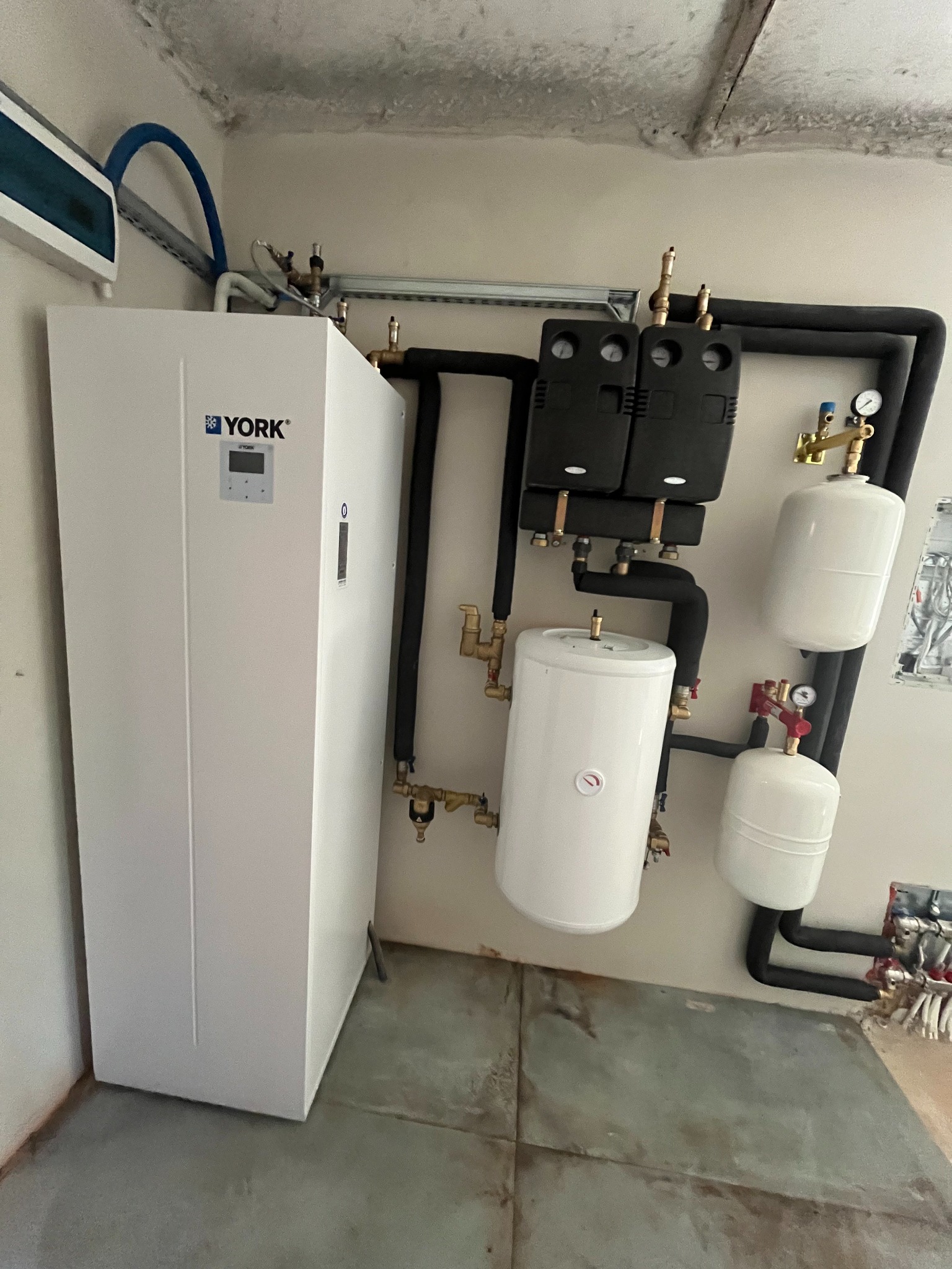 Biała jednostka wewnętrzna z logo YORK podłączona do instalacji hydraulicznej z czarnymi rurami, zbiornikami wyrównawczymi i manometrami, zamontowana w kotłowni z surowym betonowym sufitem.