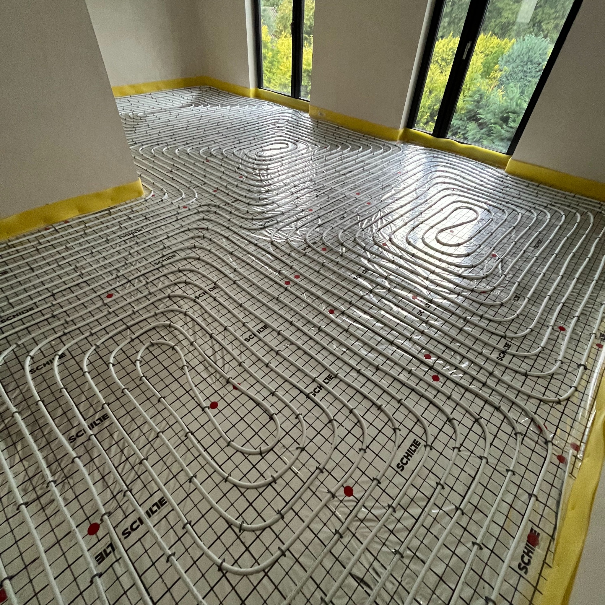 Białe rury ogrzewania podłogowego ułożone na czarnej siatce z folią, widoczne okna z zielenią za nimi i żółta taśma brzegowa.