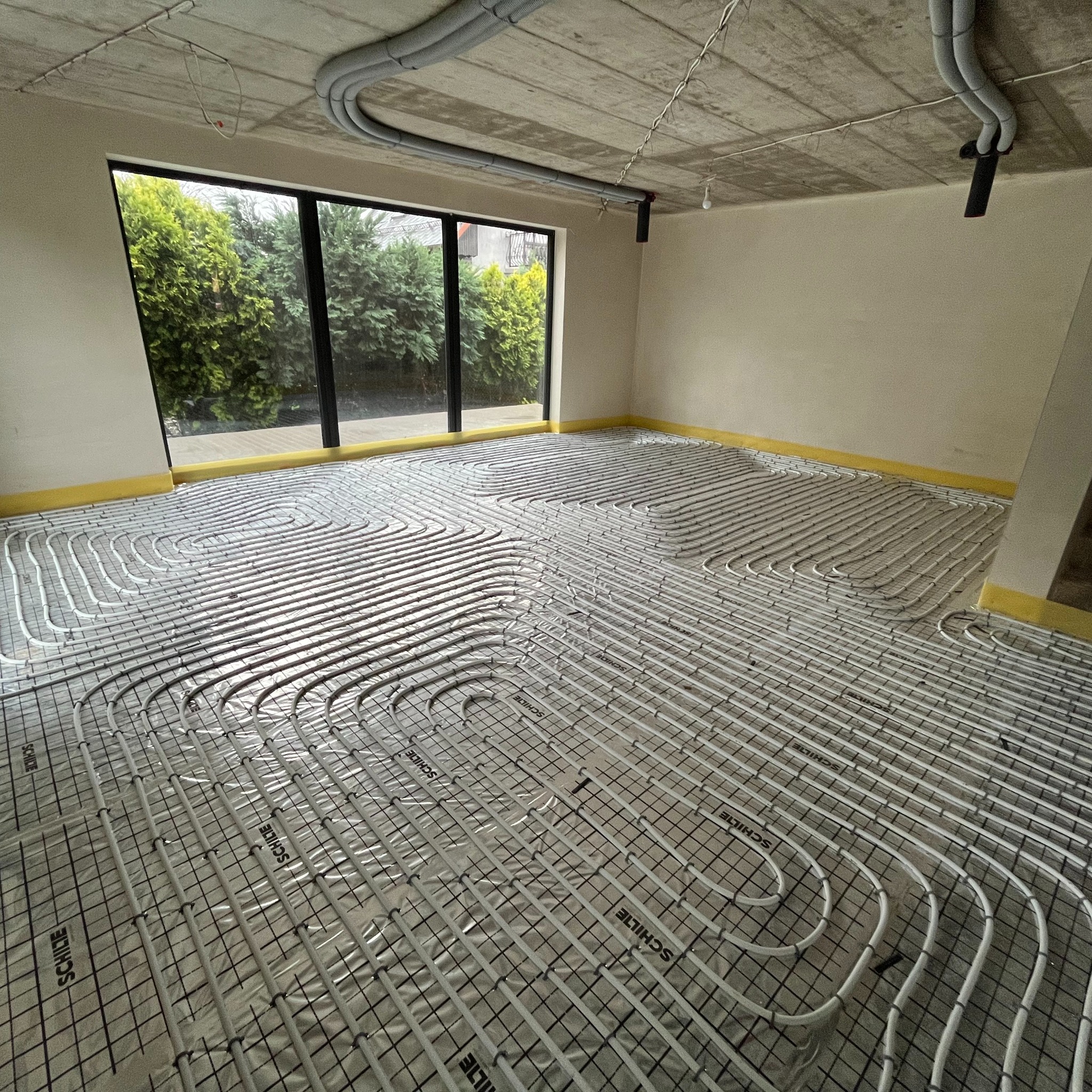 Wnętrze w trakcie montażu ogrzewania podłogowego: rury grzewcze ułożone na folii z siatką, widoczne okno z widokiem na zieleń i surowy betonowy sufit z instalacjami.