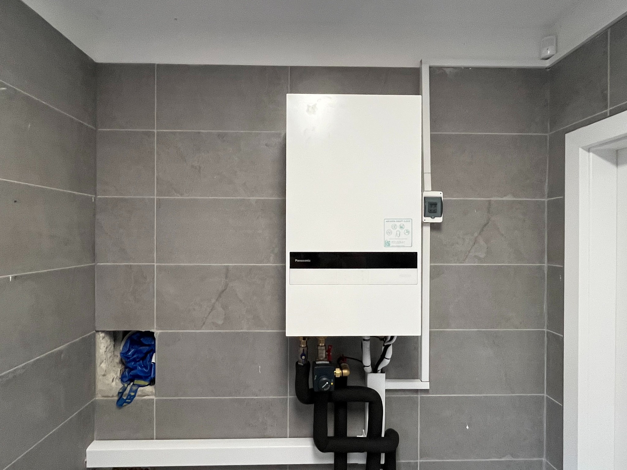 Biała jednostka wewnętrzna z czarnymi rurami instalacji grzewczej na szarej, kafelkowanej ścianie. Widoczny otwór w ścianie z niebieską folią zabezpieczającą.