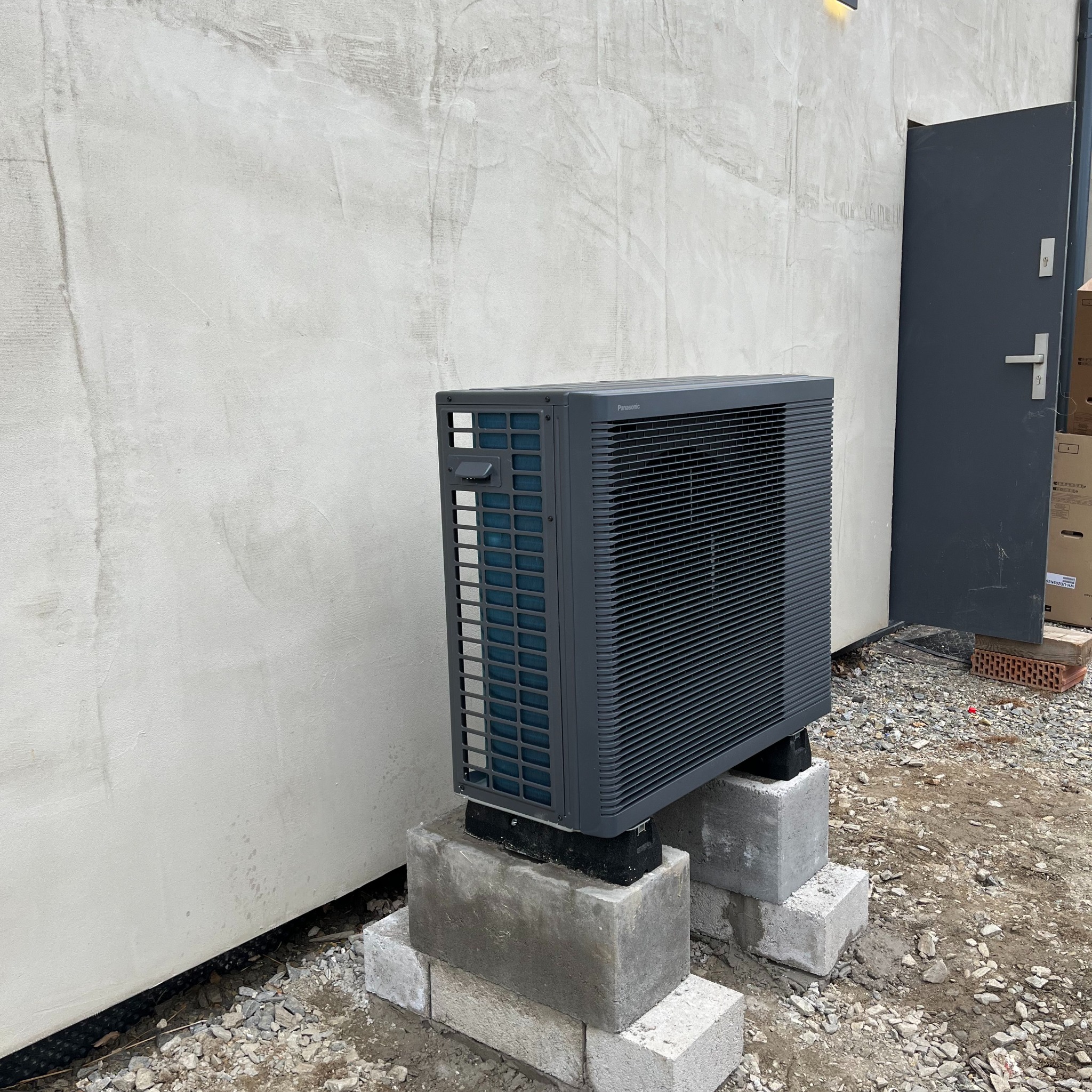 Zewnętrzna jednostka klimatyzacji lub pompy ciepła, marki Panasonic, zamontowana na betonowych bloczkach przy ścianie budynku z widocznymi śladami prac budowlanych.