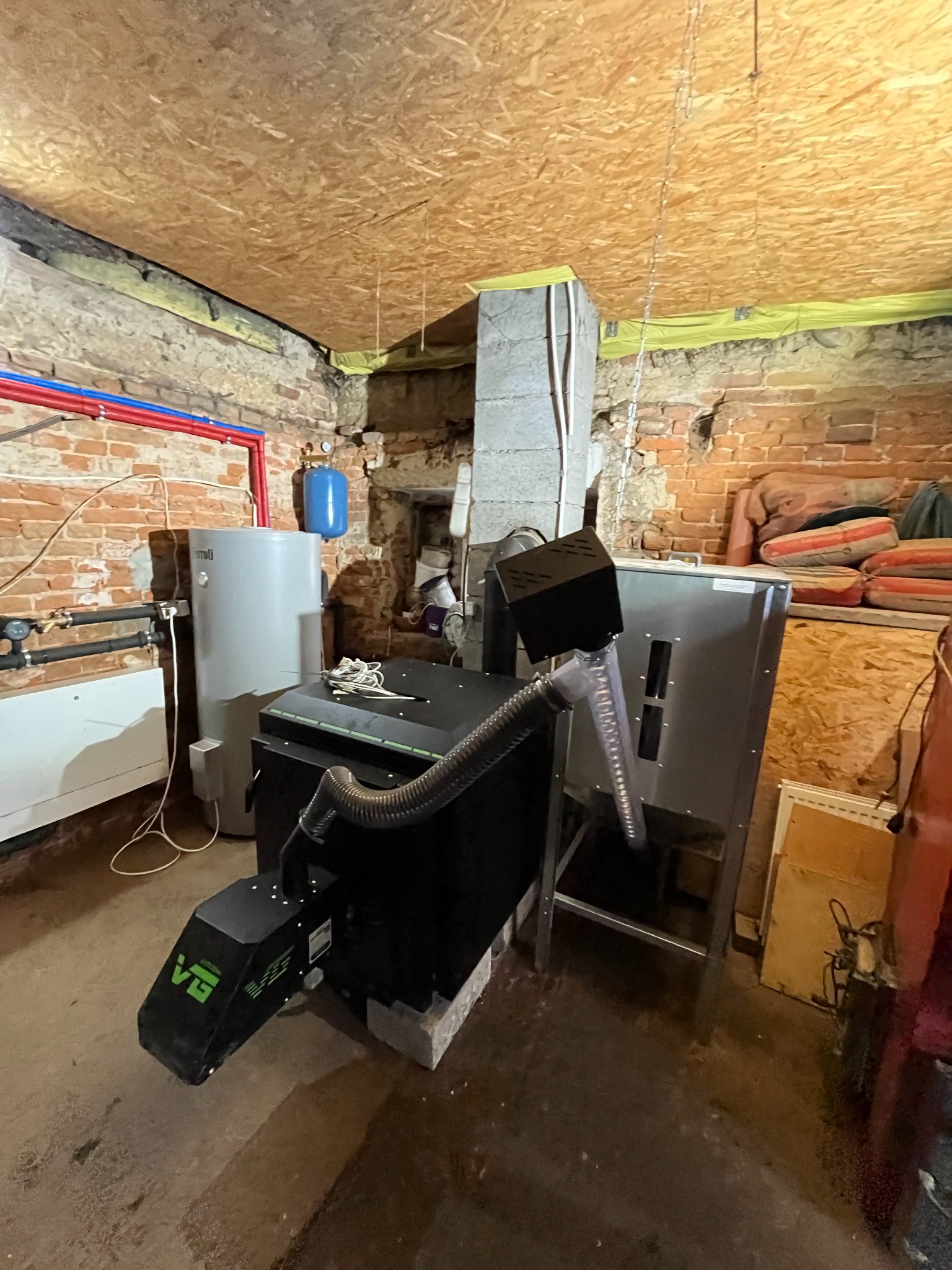 Kotłownia z piecem na paliwo stałe, zbiornikiem buforowym i instalacją hydrauliczną w piwnicy z ceglanymi ścianami i drewnianym stropem.