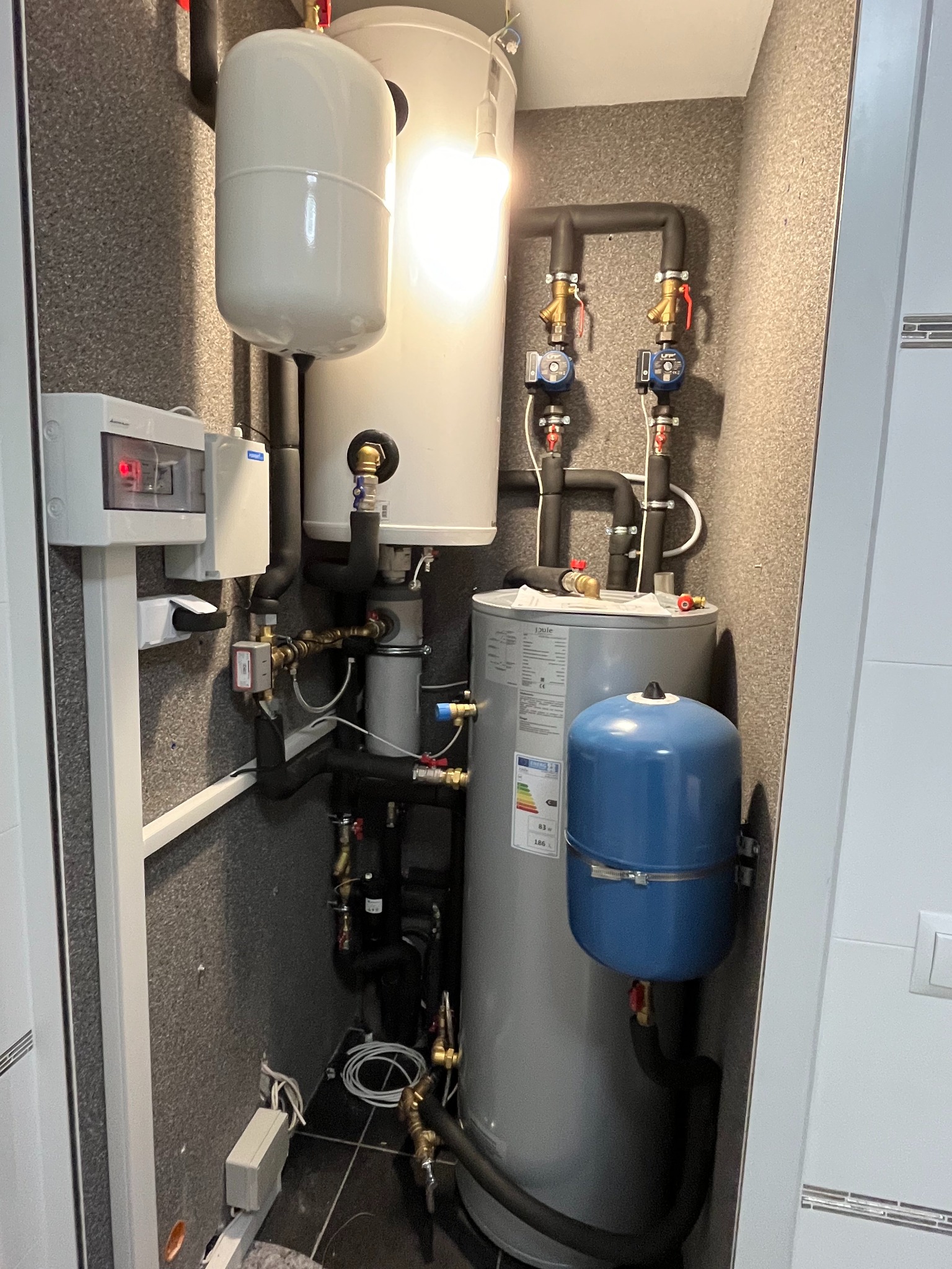 Kompaktowa instalacja grzewcza w pomieszczeniu technicznym z szarymi ścianami, obejmująca białe zbiorniki, szary bojler z niebieskim naczyniem przeponowym oraz liczne rury i zawory.