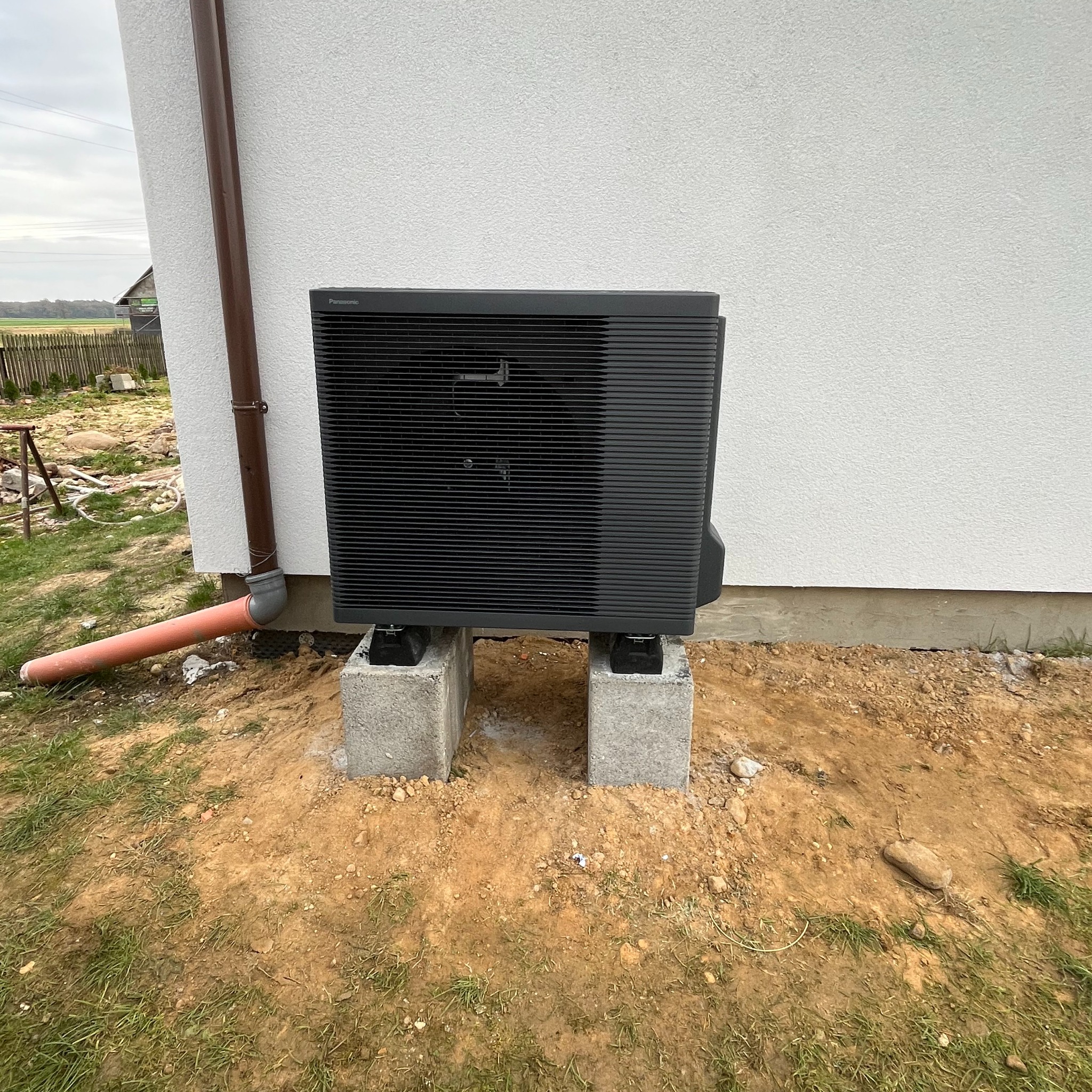 Czarna pompa ciepła Panasonic Aquarea, zamontowana na dwóch betonowych bloczkach, przy białej ścianie domu w miejscowości Węża, z widocznym odpływem kondensatu.