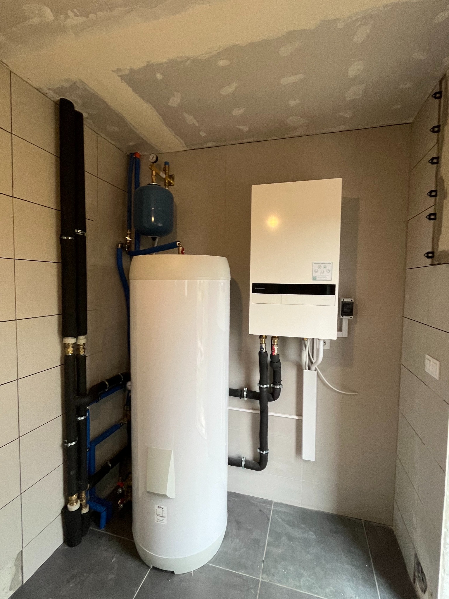 Wnętrze kotłowni z zamontowanym białym zasobnikiem ciepłej wody, niebieskim zbiornikiem wyrównawczym, oraz wiszącym białym kotłem grzewczym na ścianie z szarych płytek. Rury w izolacji termicznej...