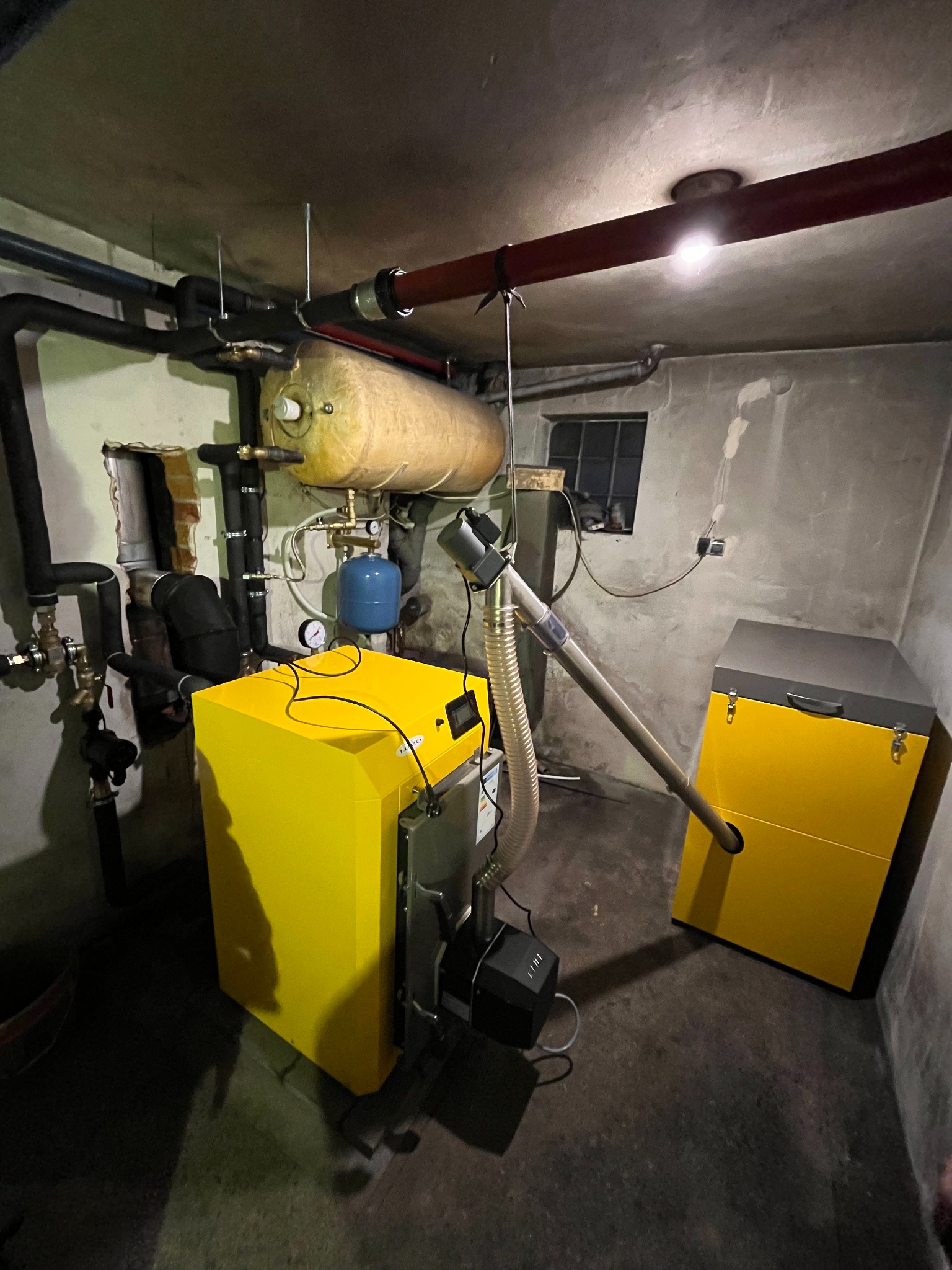 Żółty piec z podajnikiem w kotłowni, widoczne rury instalacji grzewczej i zbiornik wyrównawczy, małe okno w tle.
