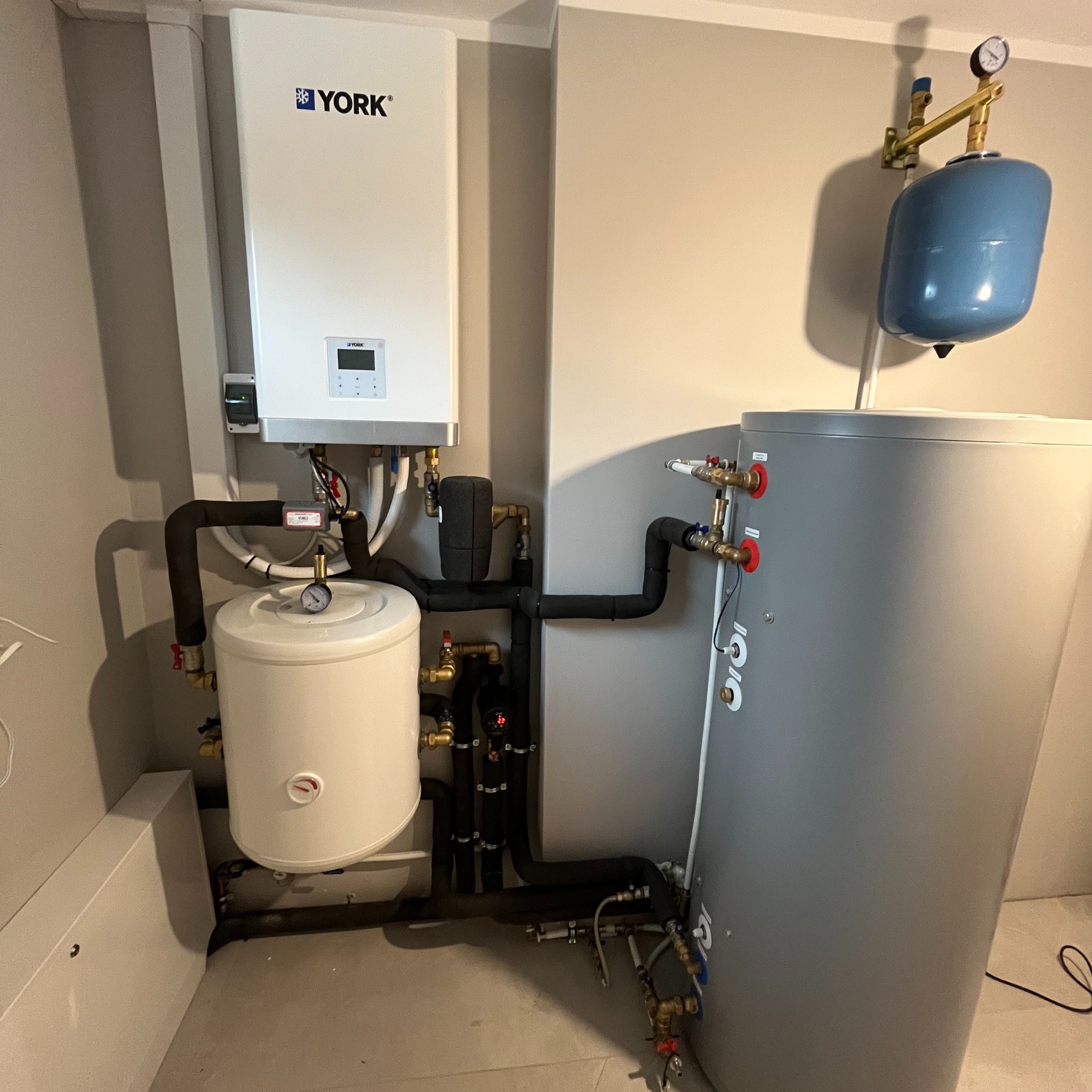 Instalacja grzewcza z białym zasobnikiem i szarym zbiornikiem buforowym, rury w izolacji, manometry, marka York na górnym urządzeniu.