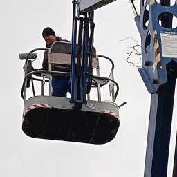 Pracownik w podnośniku koszowym, maluje elewację budynku. Widoczny fragment konstrukcji podnośnika i malarz z wałkiem. Ujęcie z dołu, pochmurne niebo w tle.