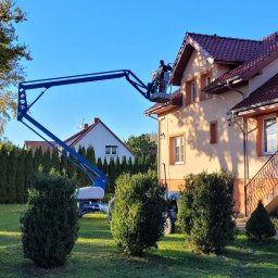 USŁUGI REMONTOWO BUDOWLANE JAROSŁAW SOSNOWSKI - Pracownik na podnośniku koszowym przy elewacji domu z czerwoną dachówką, wykonujący prace na wysokości. Widok z ogrodu, słoneczny dzień.