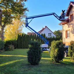 USŁUGI REMONTOWO BUDOWLANE JAROSŁAW SOSNOWSKI - Praca na wysokościach: Dwóch pracowników na podnośniku koszowym przy dachu domu jednorodzinnego w słoneczny dzień. Widok z ogrodu, zieleń w tle.