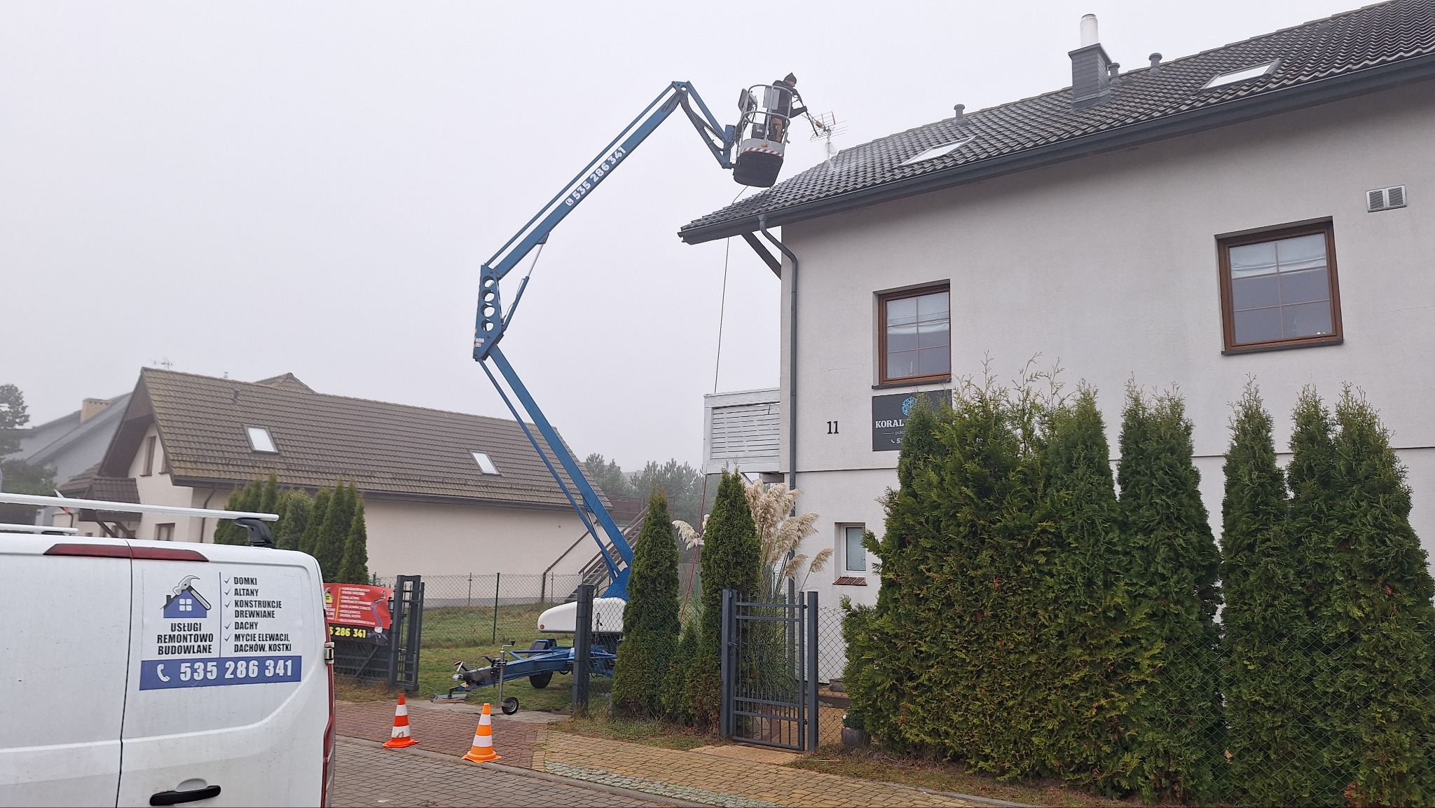 Pracownik na podnośniku teleskopowym przy dachu domu jednorodzinnego. W tle zabudowa osiedlowa i firmowy bus z numerem telefonu.