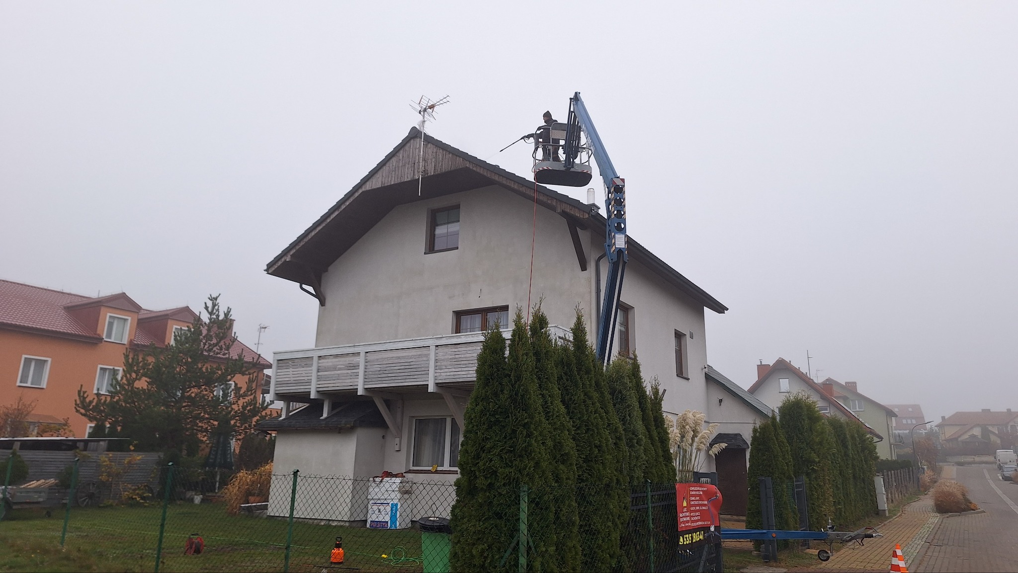 Pracownik na podnośniku czyści dach domu jednorodzinnego z anteną, używając myjki ciśnieniowej. Widoczne drzewa iglaste i zabudowa mieszkalna w tle, pochmurny dzień.