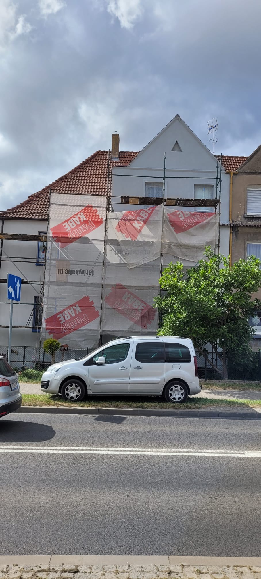 Dom w rusztowaniach z siatką reklamową Farby Kabe, widok z ulicy. Biały samochód zaparkowany przed budynkiem. Częściowo widoczny dach z czerwonej dachówki.