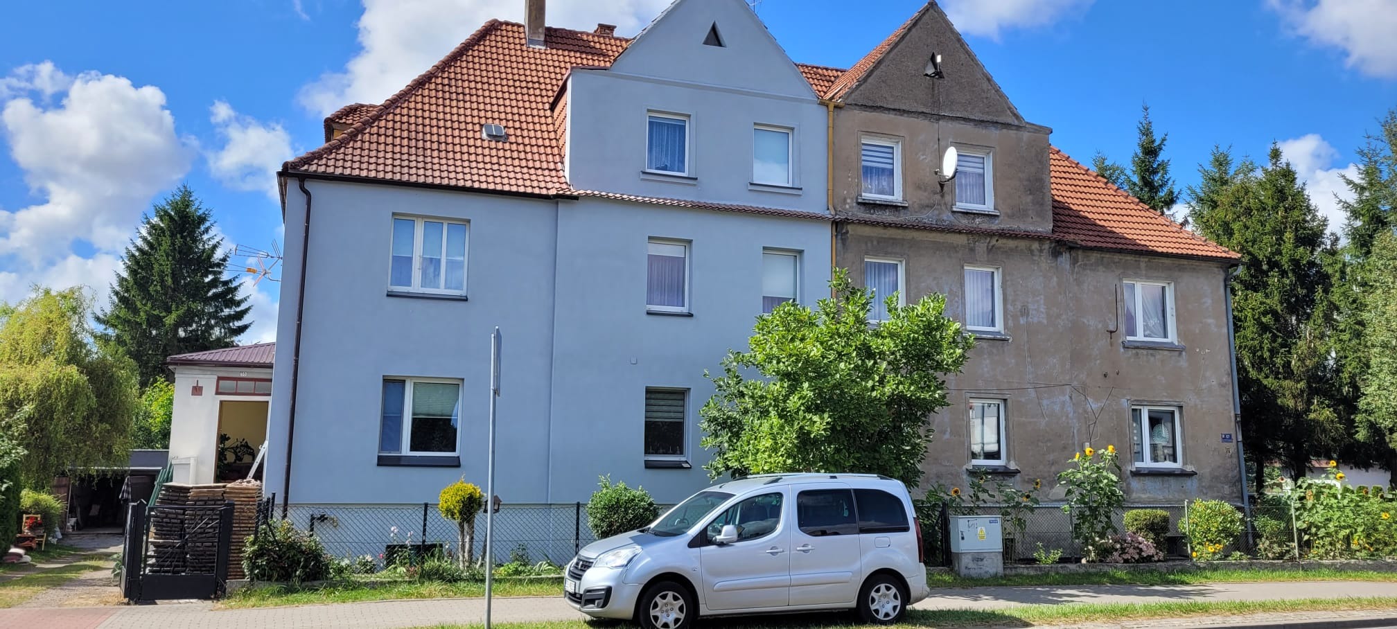 Budynek po ociepleniu elewacji, z widoczną różnicą w wyglądzie w porównaniu do sąsiedniej, nieocieplonej części. Czerwony dach, jasnoniebieska elewacja, srebrny samochód.