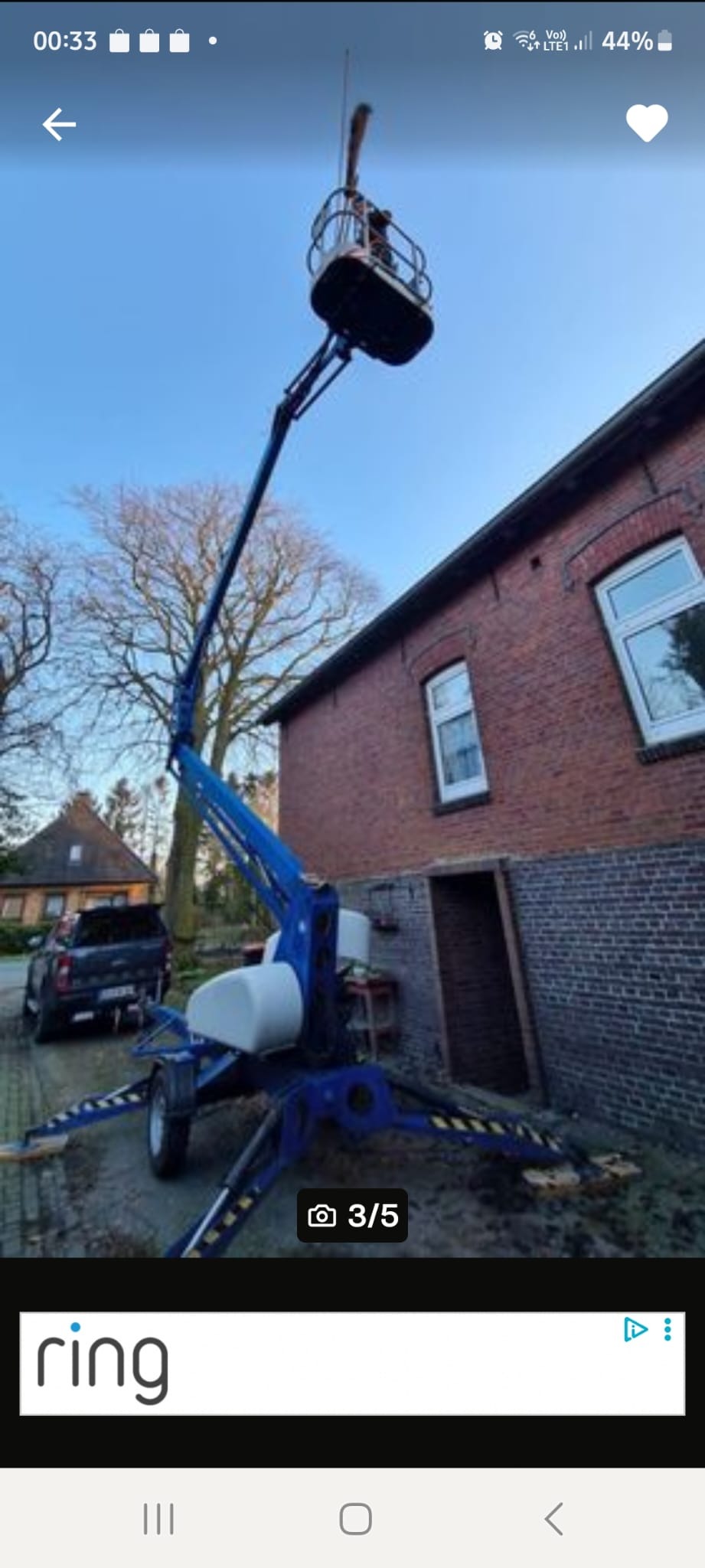 Niebieski podnośnik koszowy na tle ceglanego budynku i drzew bez liści. Osoba w koszu. Widoczne podpory i koła podnośnika. Błękitne niebo w tle.