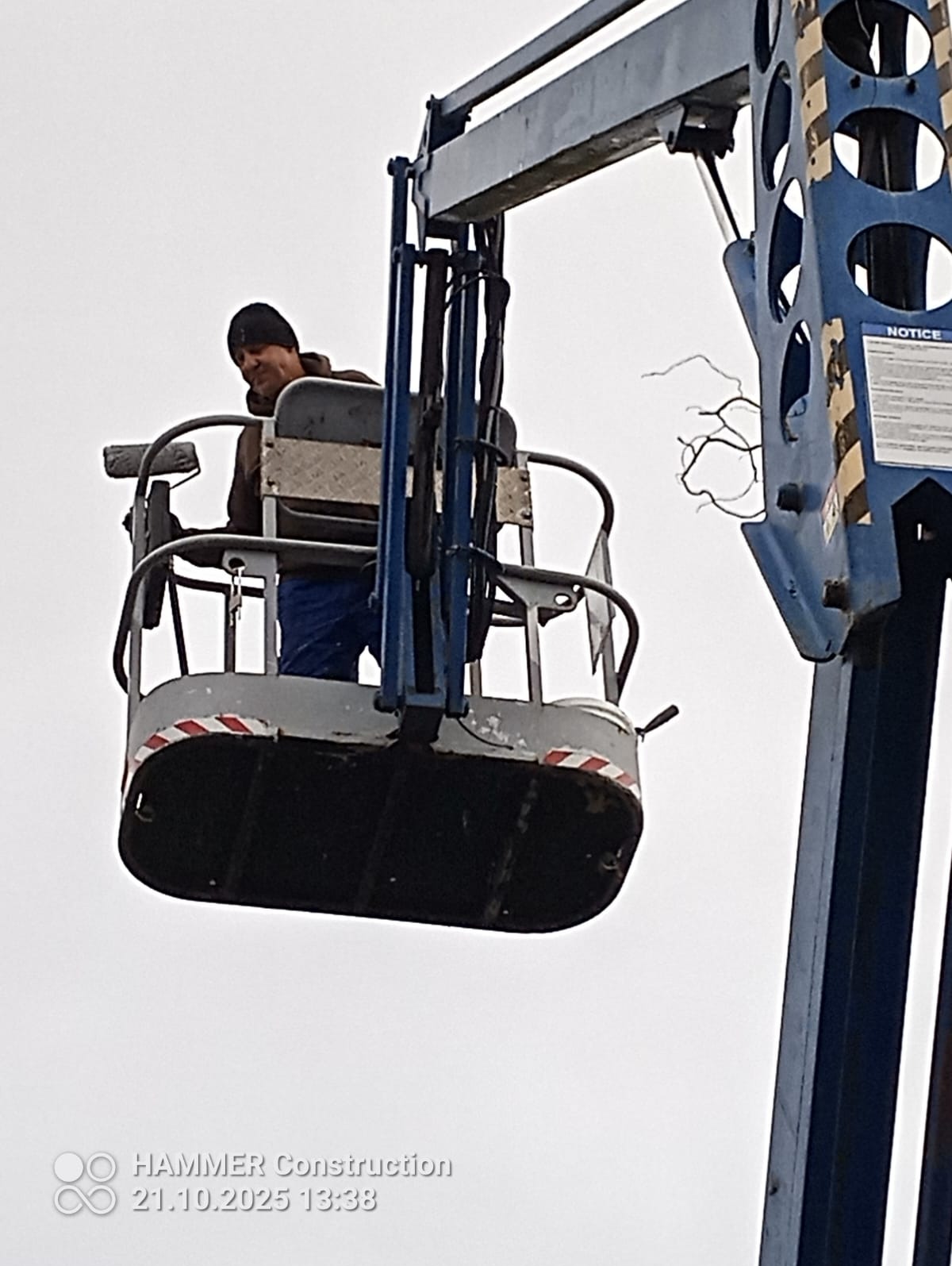 Pracownik w podnośniku koszowym, maluje elewację budynku. Widoczny fragment konstrukcji podnośnika i malarz z wałkiem. Ujęcie z dołu, pochmurne niebo w tle.