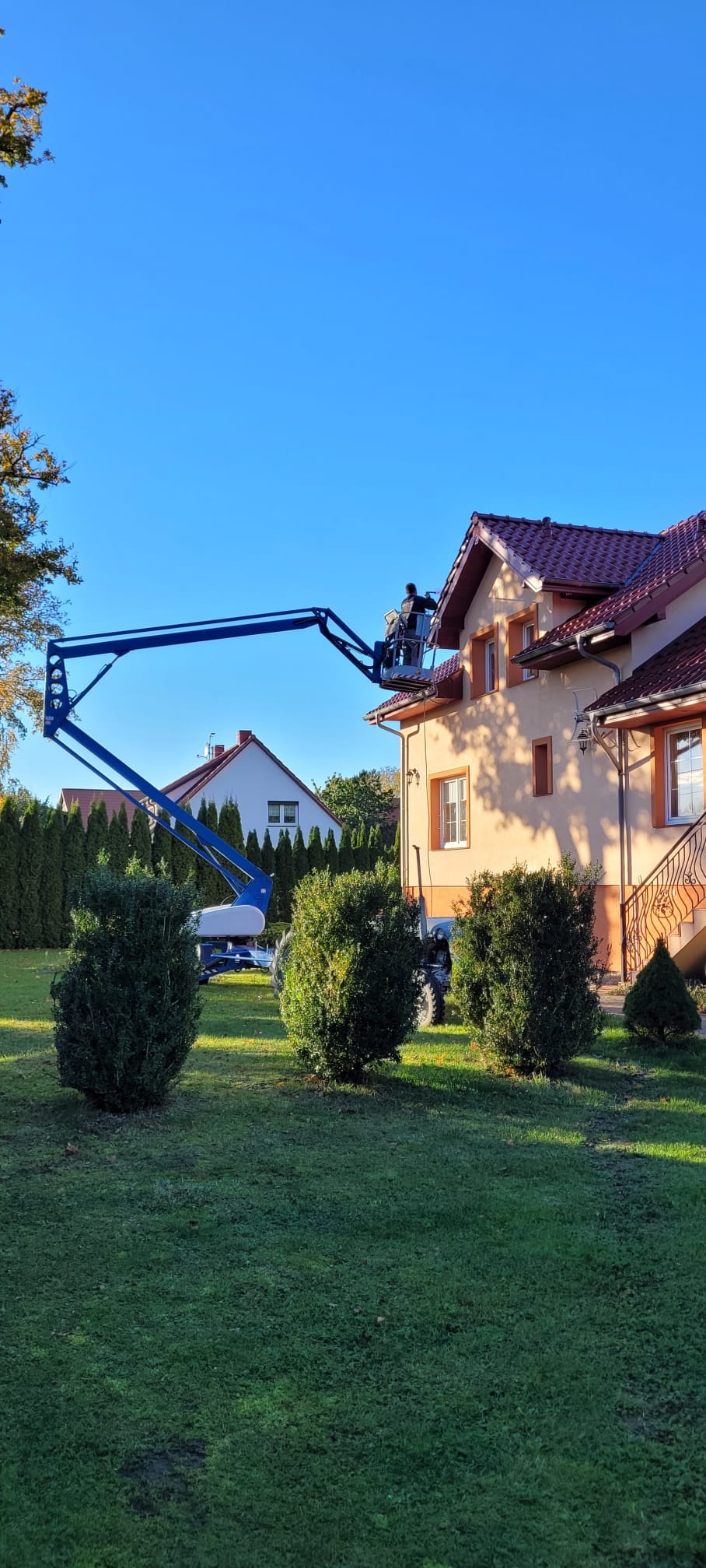 Pracownik na podnośniku koszowym przy elewacji domu z czerwoną dachówką, wykonujący prace na wysokości. Widok z ogrodu, słoneczny dzień.