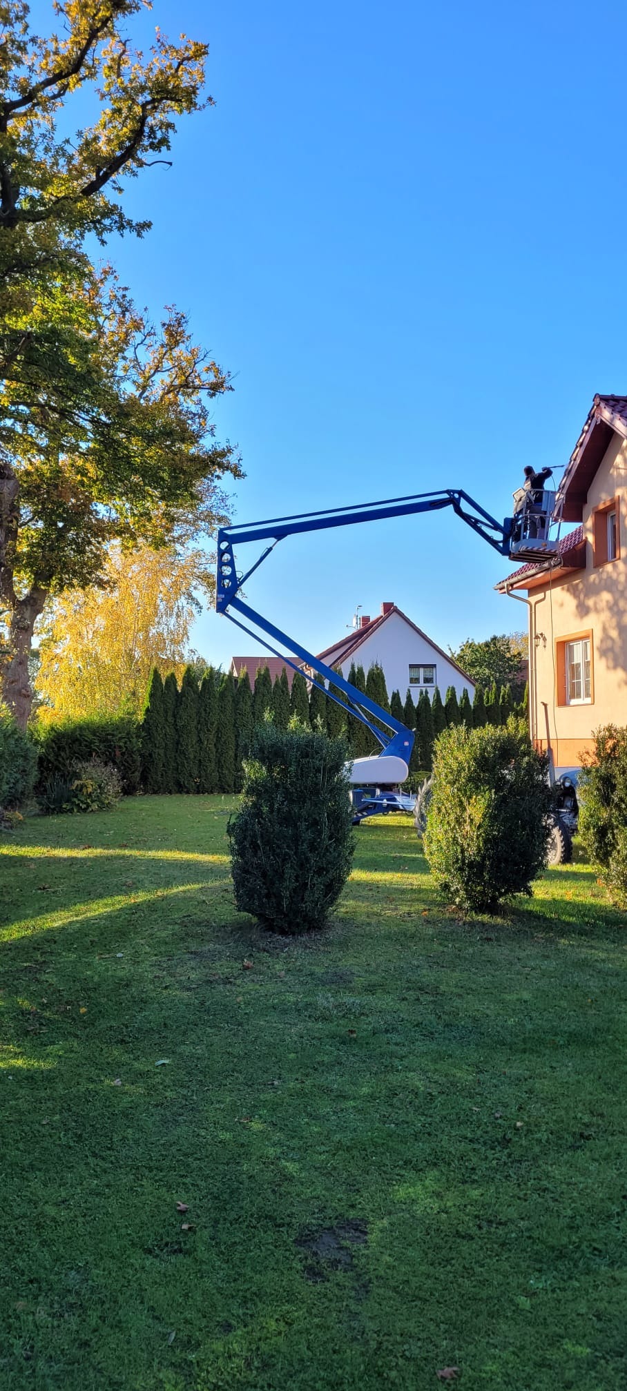 Praca na wysokościach: Dwóch pracowników na podnośniku koszowym przy dachu domu jednorodzinnego w słoneczny dzień. Widok z ogrodu, zieleń w tle.