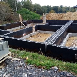 BM TWÓJ DOM SPÓŁKA Z OGRANICZONĄ ODPOWIEDZIALNOŚCIĄ - Wykopane fundamenty nowego domu z bloczków betonowych, zabezpieczone czarną izolacją, widoczne zbrojenia i instalacje, w tle ziemia i zieleń.