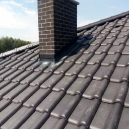 Ciemna dachówka ceramiczna z kominem z cegły klinkierowej na tle błękitnego nieba z chmurami.