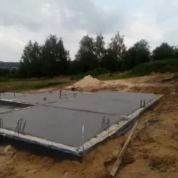 Zalany, świeży fundament płyty domu jednorodzinnego z wystającymi prętami zbrojeniowymi, zabezpieczony folią kubełkową, w tle drzewa i hałda piasku.