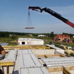 Montaż prefabrykowanych stropów na budowie domu jednorodzinnego przy użyciu dźwigu, w tle widoczny budynek w trakcie wznoszenia ścian z czerwonej cegły.