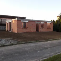 Surowy stan domu z czerwonej cegły, widoczne otwory okienne i drzwiowe, szary fundament, przygotowany teren pod budowę. Dom w trakcie wznoszenia ścian.