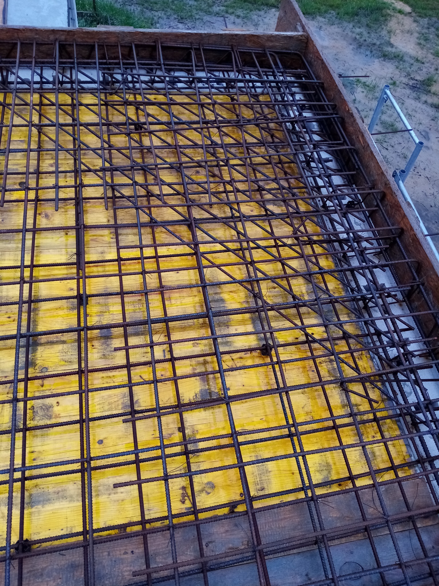Zbrojenie fundamentu domu z widoczną żółtą szalunką i drewnianą ramą, przygotowane do zalania betonem.